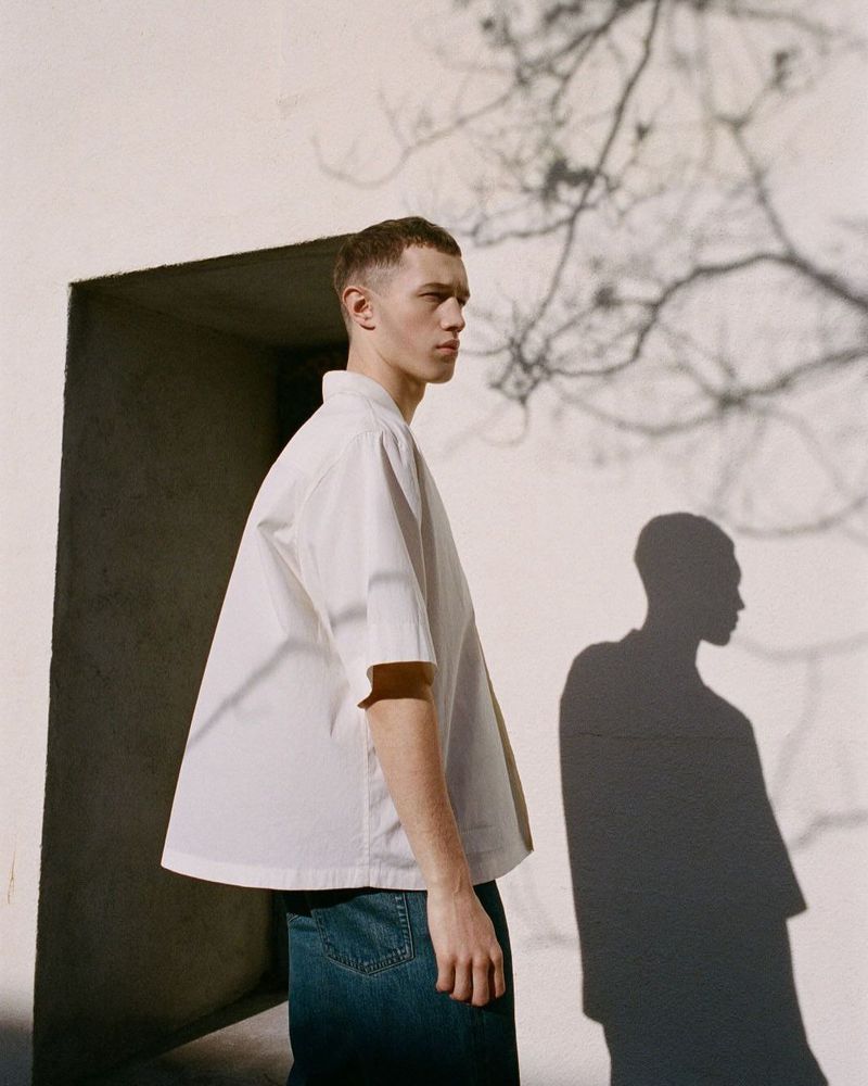 Young man wearing a white short-sleeve shirt and denim jeans standing outdoors against a plain wall with tree shadows.