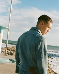 Man wearing a blue denim jacket standing near a rocky coast with ocean waves and a clear sky