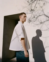 Young man wearing a white short-sleeve shirt and denim jeans standing outdoors against a plain wall with tree shadows.