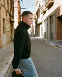 A young man wearing a black jacket and light wash jeans standing on a narrow sunny city street, holding a skateboard.