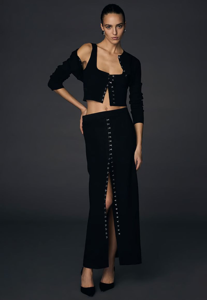 Elegant woman in a black knitted crop top, cardigan, and maxi skirt with slit poses in a studio setting, showcasing fashion apparel.