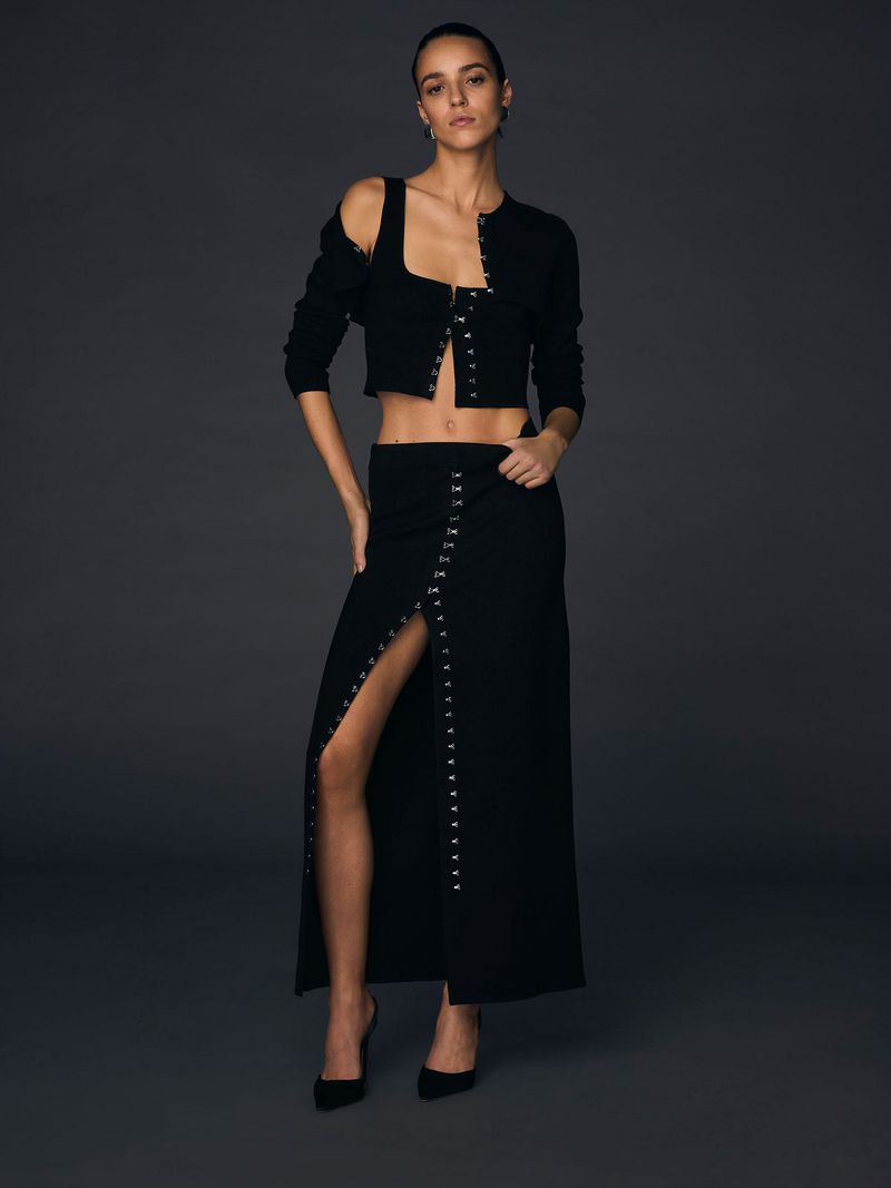 Woman poses in a black, two-piece skirt set with black high heels against a grey studio backdrop for a fashion editorial shot.