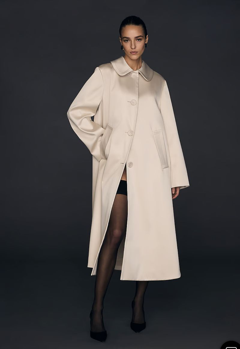 A woman wearing a cream-colored trench coat, black stockings, and heels poses in a studio with soft, neutral lighting.