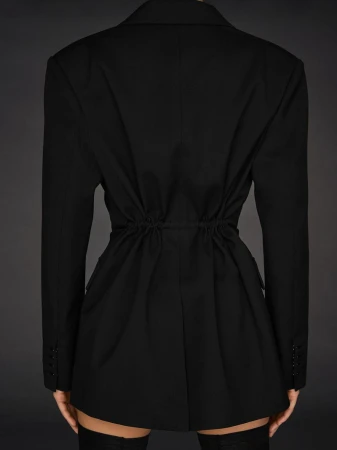 Back view of woman wearing a black blazer dress and high black boots standing against a gray background in a studio environment.
