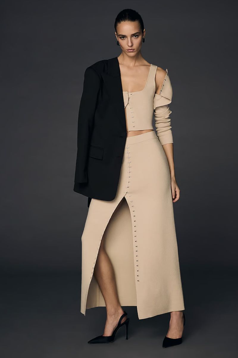 Fashionable woman in beige two-piece dress with black blazer, posing confidently in a studio with a smooth grey backdrop.