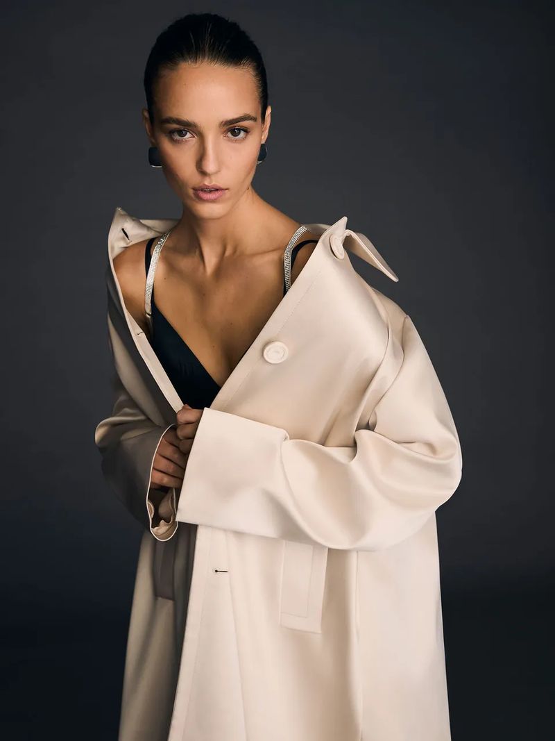 A woman in a beige coat stands against a grey background looking at the camera. Her dark hair is pulled back and she wears black earrings.