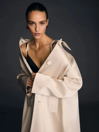 A woman in a beige coat stands against a grey background looking at the camera. Her dark hair is pulled back and she wears black earrings.