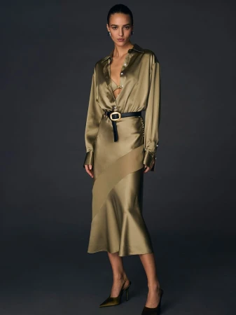 Model Emm Arruda wearing a glossy olive satin blouse and matching skirt with a black belt against a dark grey background.