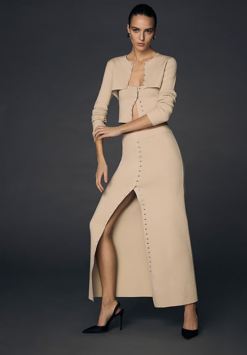 Model Emm Arruda wearing a beige knit two-piece set with a button-front skirt against a dark grey seamless studio background.