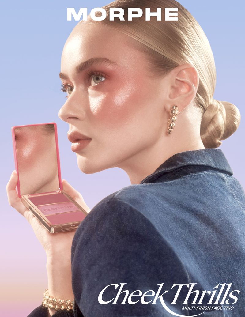 Blonde woman showcasing Morphe blush palette; pink hues highlight cheeks, complemented by a denim top against a soft blue backdrop.