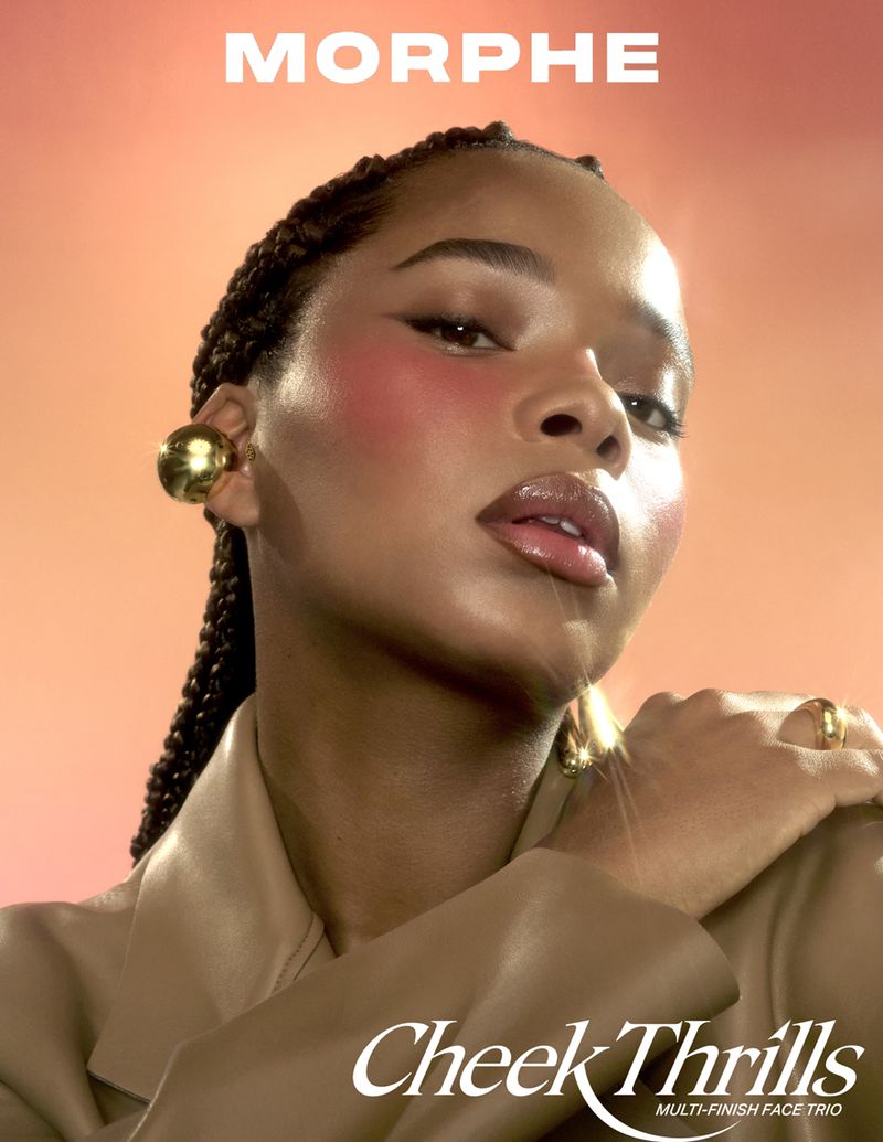 Beauty shot of a Black woman with braided hair wearing blush and a tan jacket in front of a coral gradient background, advertising makeup.