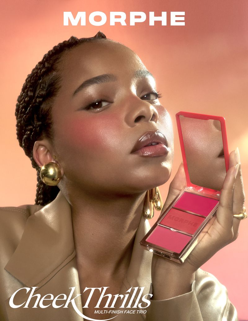 A woman with braids is holding a makeup palette. She has on gold earrings, a beige jacket, and blush on her cheeks, on peach background.