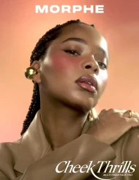 Close-up portrait of a black woman wearing bold blush, gold jewelry, and a camel jacket against a warm gradient background.